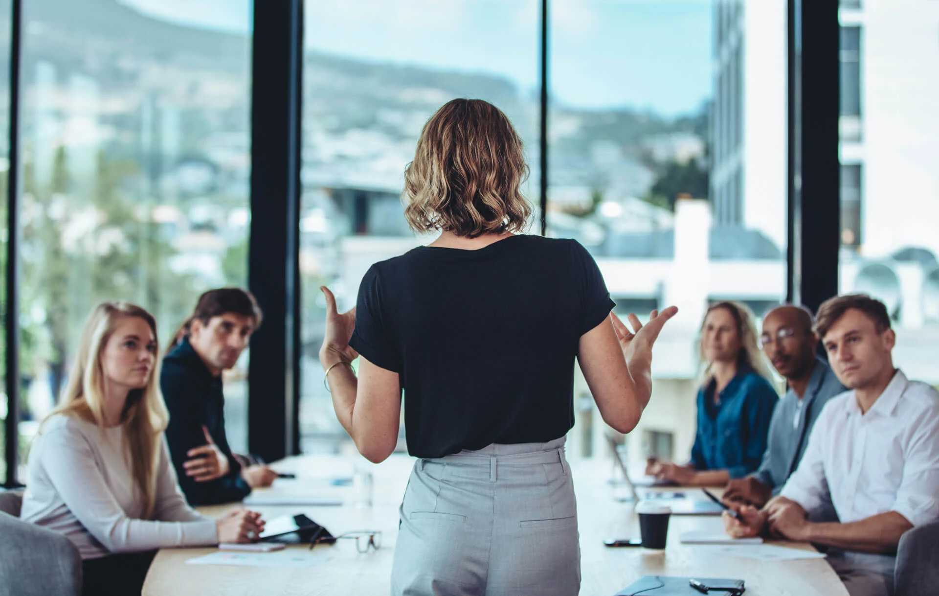 A woman stands and speaks to a group of attentive colleagues seated around a conference table featuring sustainable surfaces in a modern office with large windows and a cityscape view.