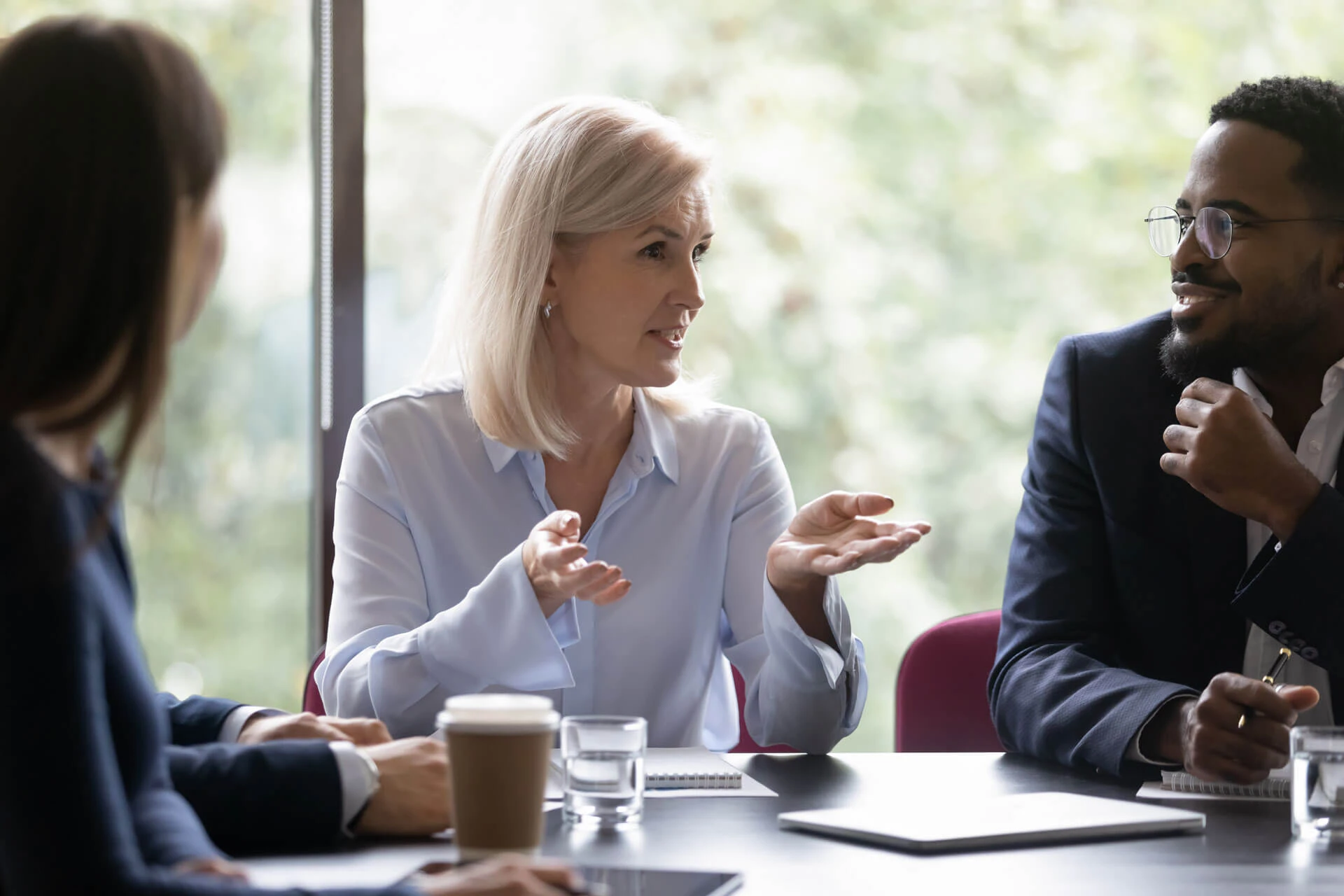 A woman with blonde hair gestures while speaking to colleagues during a meeting at a table featuring sustainable surfaces, with notebooks, papers, water glasses, and coffee cups. Other people listen attentively as greenery is visible through the window.