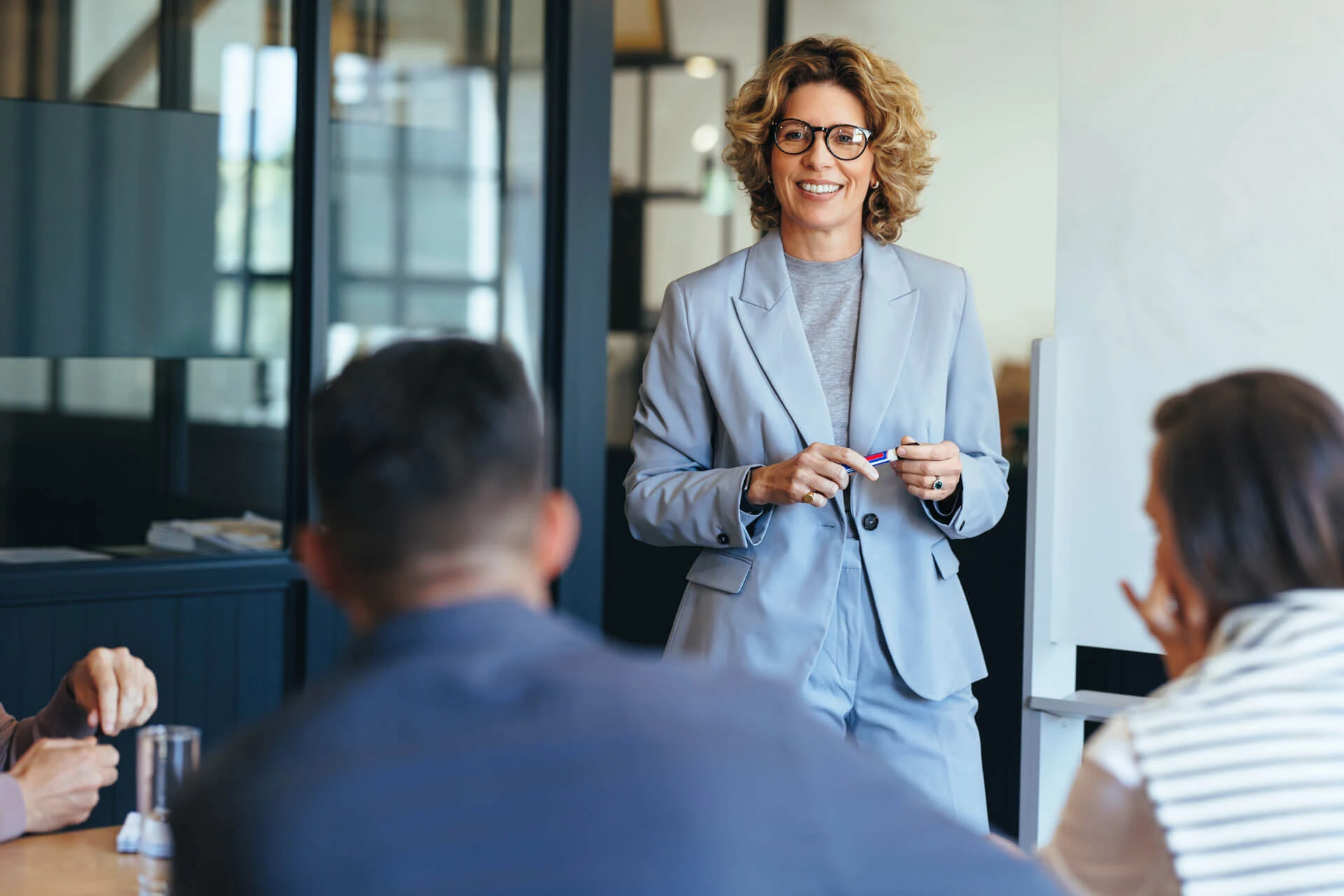 A woman in a light gray suit smiles while standing and speaking to three people seated at a conference table in a modern office setting.