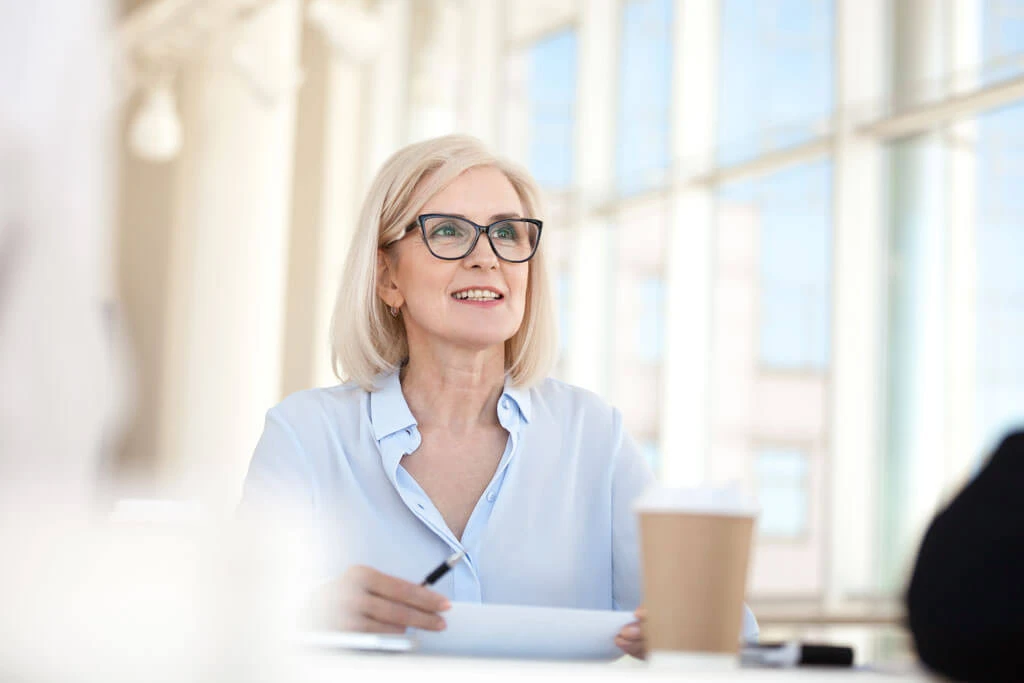A smiling older woman with blonde hair and glasses sits at a desk holding a pen, with paper and a coffee cup in front of her, surrounded by forward-thinking materials in a bright, modern office space with large windows.