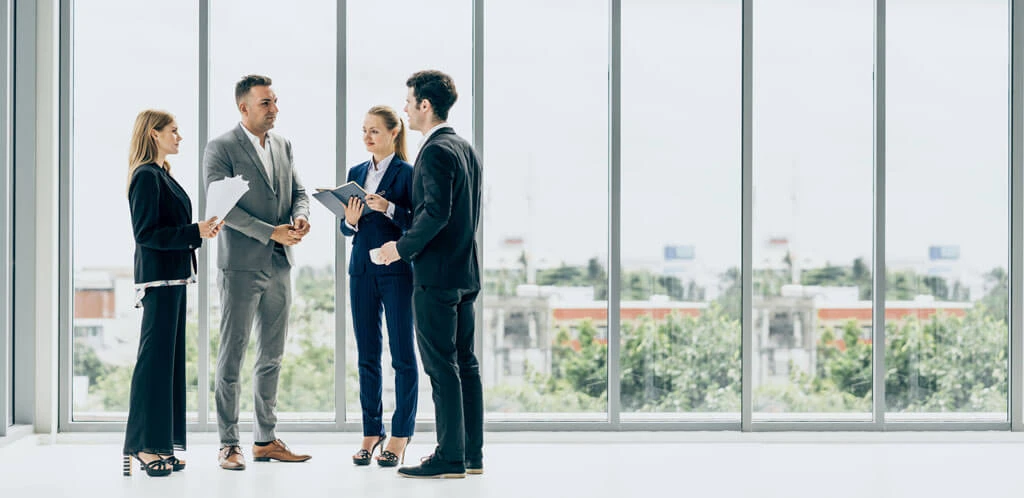 Four business professionals stand in a modern office by large windows, engaged in conversation and holding documents made from forward-thinking materials, with a cityscape visible outside.
