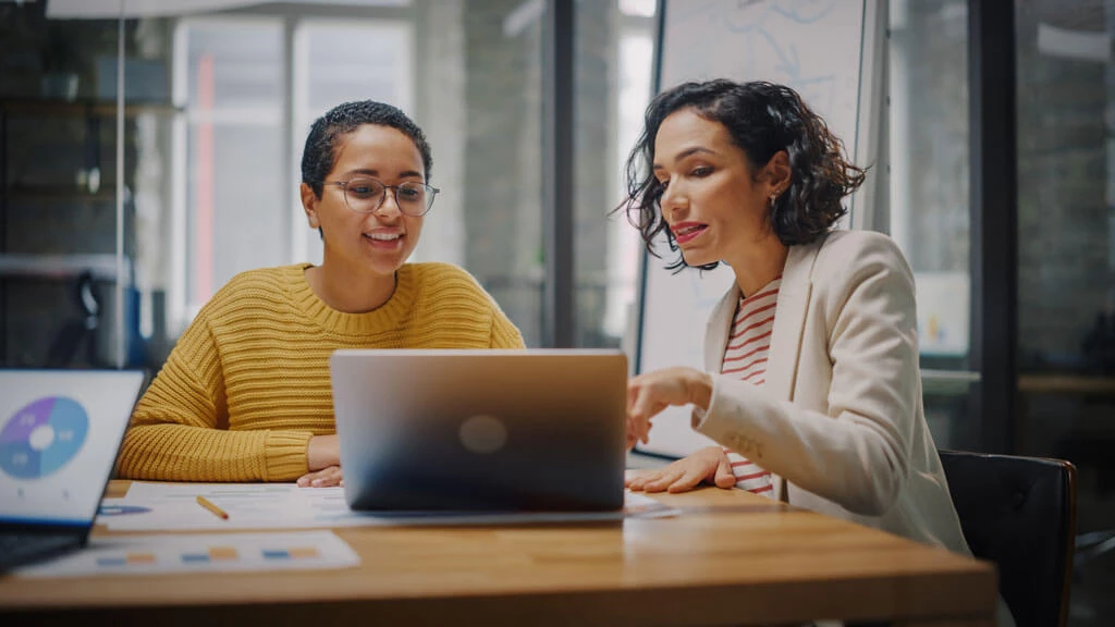 Two women sit at a desk in an office, looking at a laptop screen together. One woman points at the laptop while the other smiles, surrounded by papers and charts featuring forward-thinking materials.