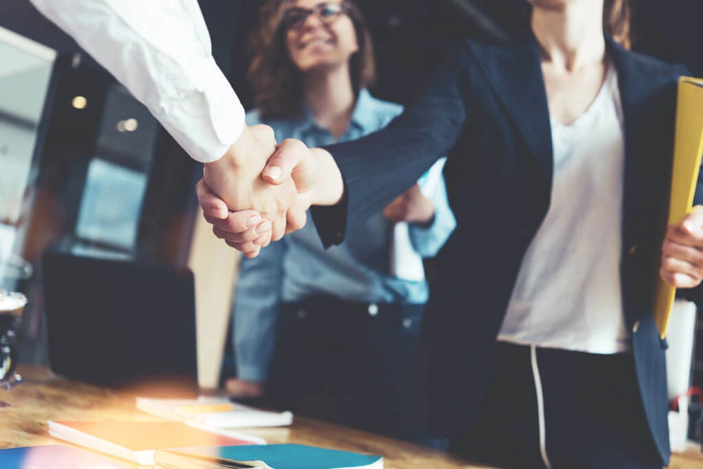 Two people in business attire shake hands over a desk with documents, as a third person smiles and watches. The professional office features forward-thinking materials, reflecting an innovative and modern work environment.