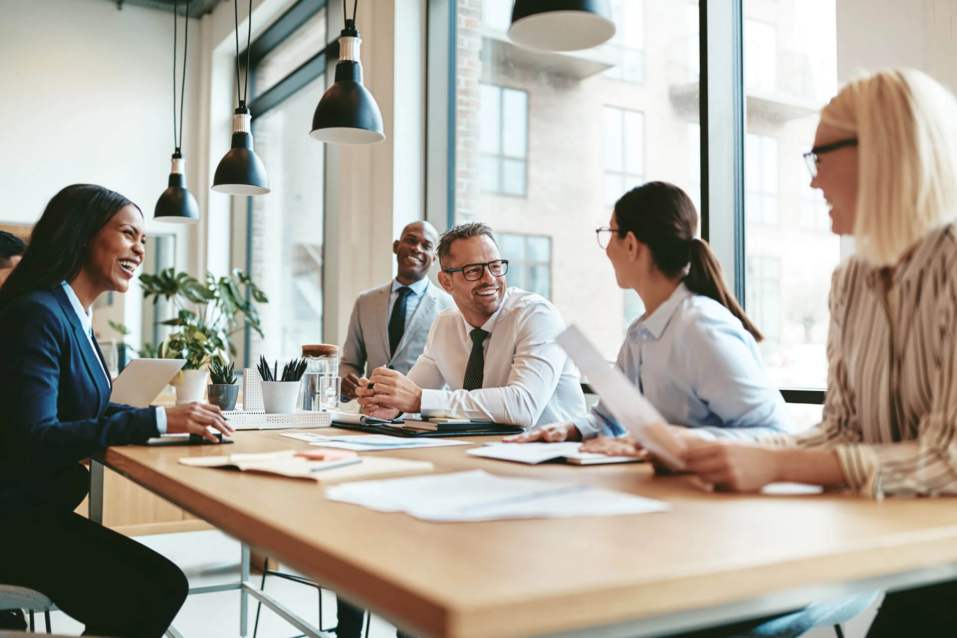Five professionally dressed people sit around a conference table in a bright, modern office, engaging in a lively discussion and smiling. Papers, pens, coffee cups, and forward-thinking materials are on the table, with large windows in the background.