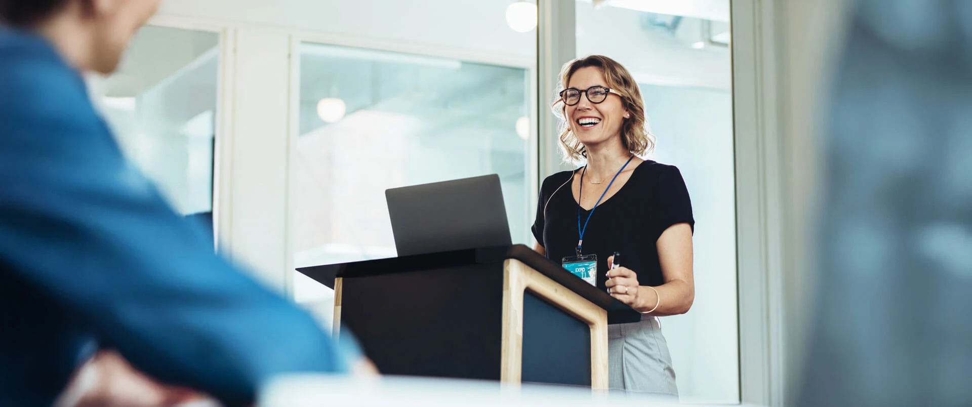 A woman wearing glasses stands at a podium with a laptop, smiling while giving a presentation in a modern, bright conference room.