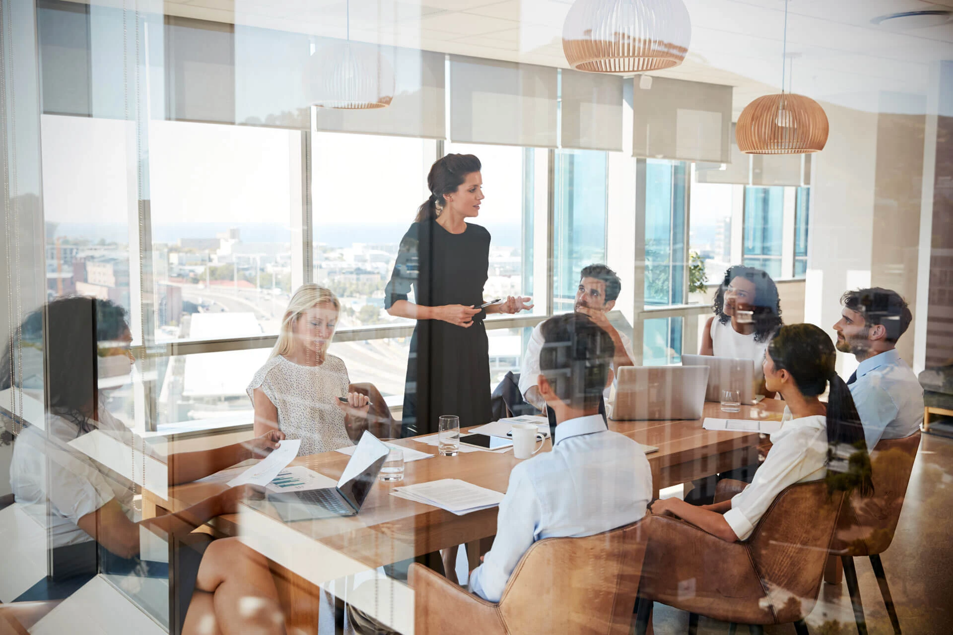 A woman stands and speaks to a group of six colleagues seated around a conference table in a bright modern office with large windows and city views. Laptops, papers, and glasses of water are on the table.