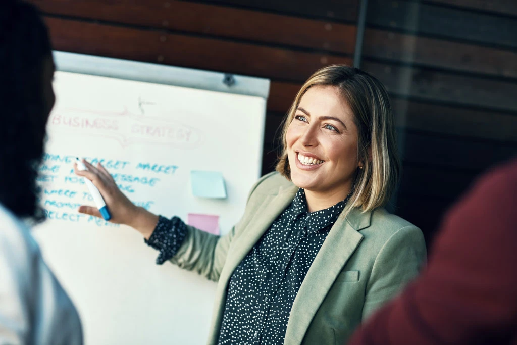 A woman in a blazer smiles while giving a presentation, gesturing at a whiteboard with notes and sticky notes. She appears engaged and is speaking to a small group in a modern office setting.