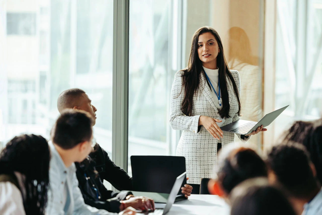 A woman in business attire stands holding a laptop and speaks to a group of people seated around a conference table in a modern office with large windows.