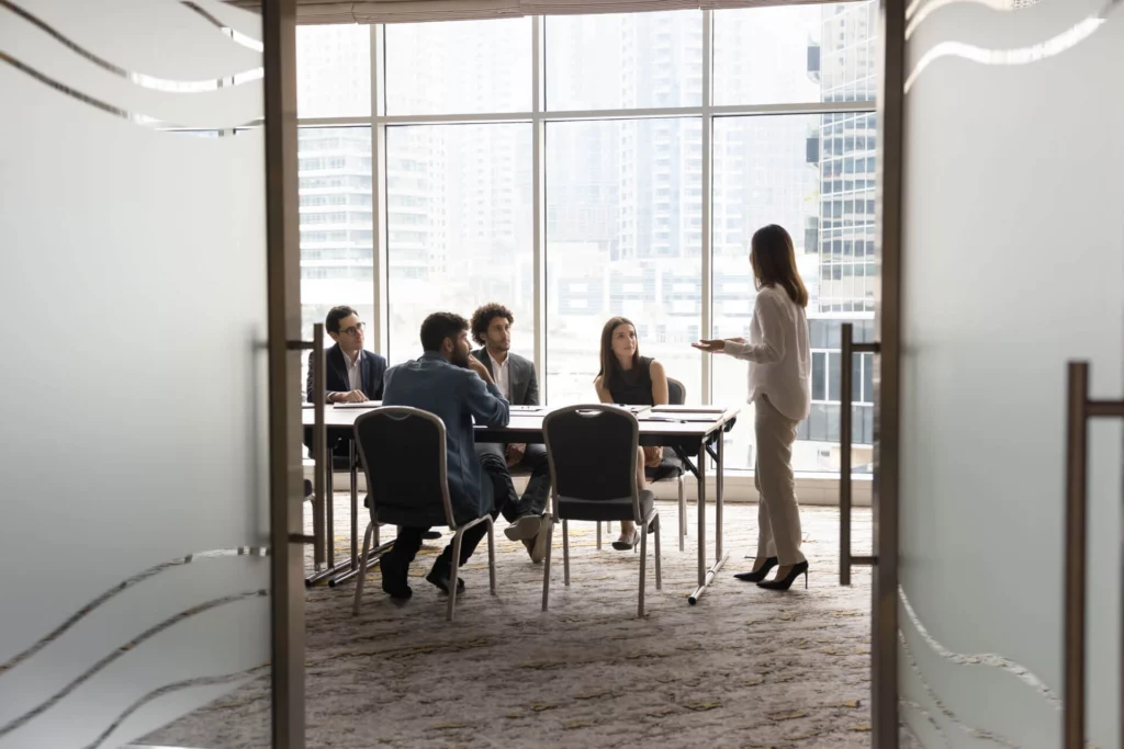 A woman stands and presents to a group of five people seated at a conference table in a modern office with large windows and a city view.