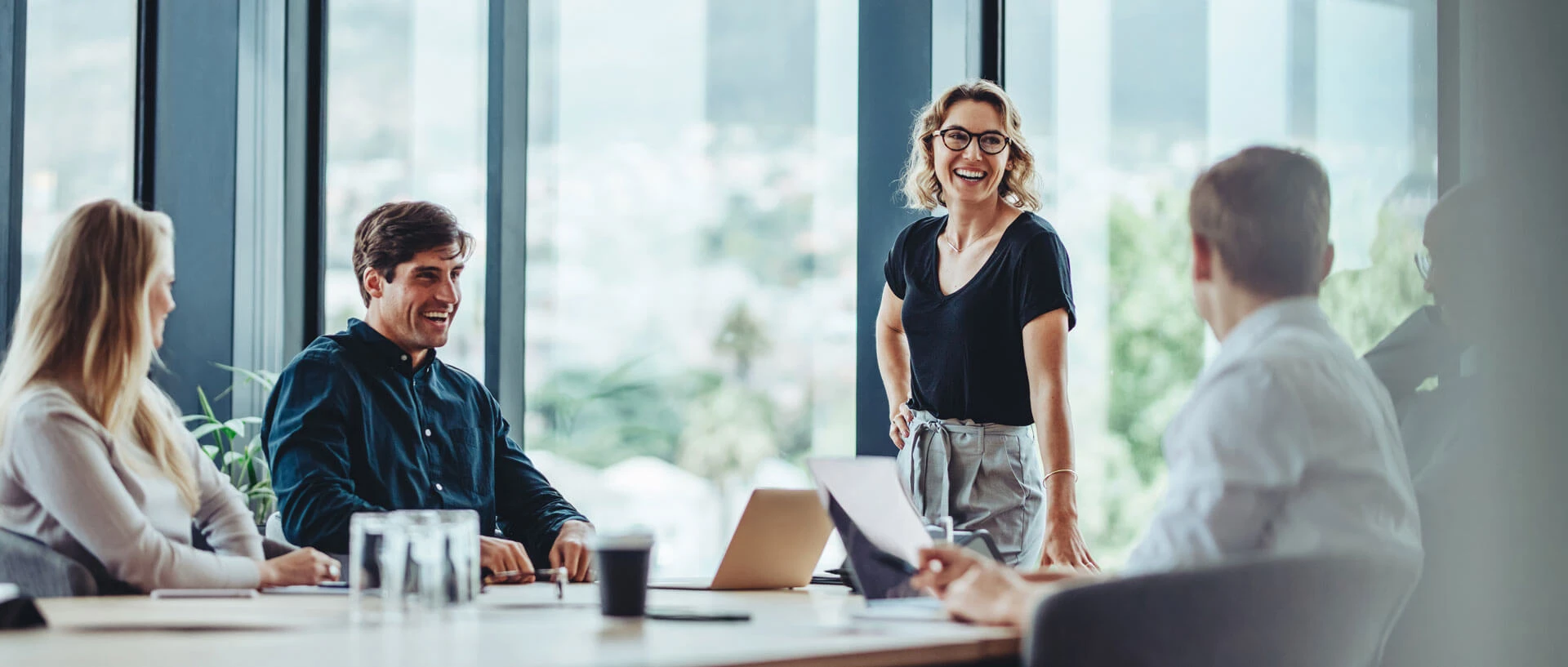 Four people are sitting at a conference table in a modern office built with 90% recycled materials. One woman stands and smiles, while the others are seated, listening and interacting with laptops and notepads.