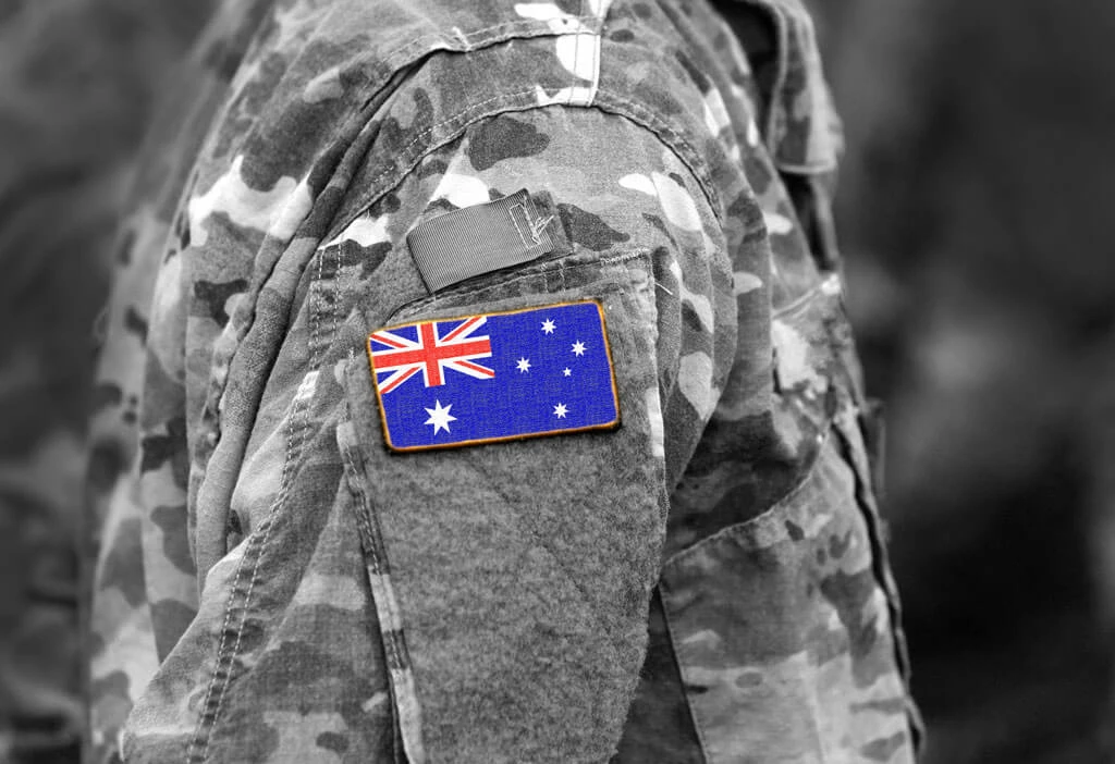 A close-up of a camouflage military uniform sleeve made from 90% recycled materials displays an Australian flag patch in color, while the rest of the image is in black and white.