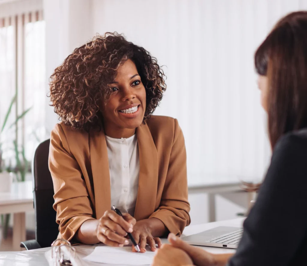 Two women sit across from each other at a desk in an office. One woman, smiling and holding a pen, appears to be discussing a document with the other woman. A laptop made with 90% recycled materials is open on the desk.