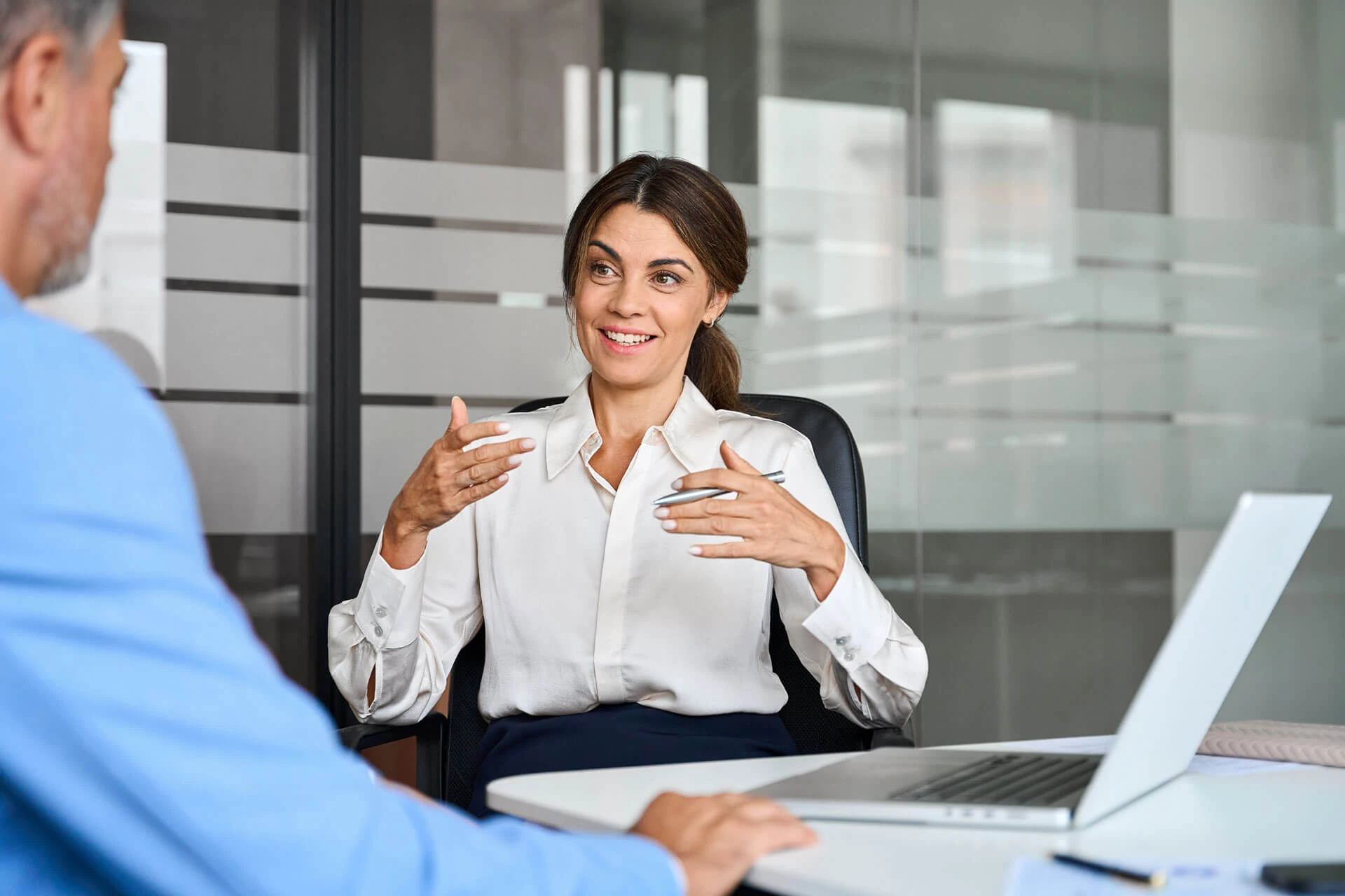 A woman in a white blouse sits at a desk talking and gesturing with her hands, while a man listens. They are in a modern office with glass walls and a laptop on the table.