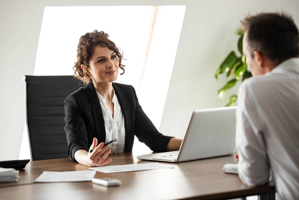 A woman in business attire sits at a desk with a laptop, smiling and talking to a man across from her. Papers and a phone are on the desk, and a plant is visible in the background.