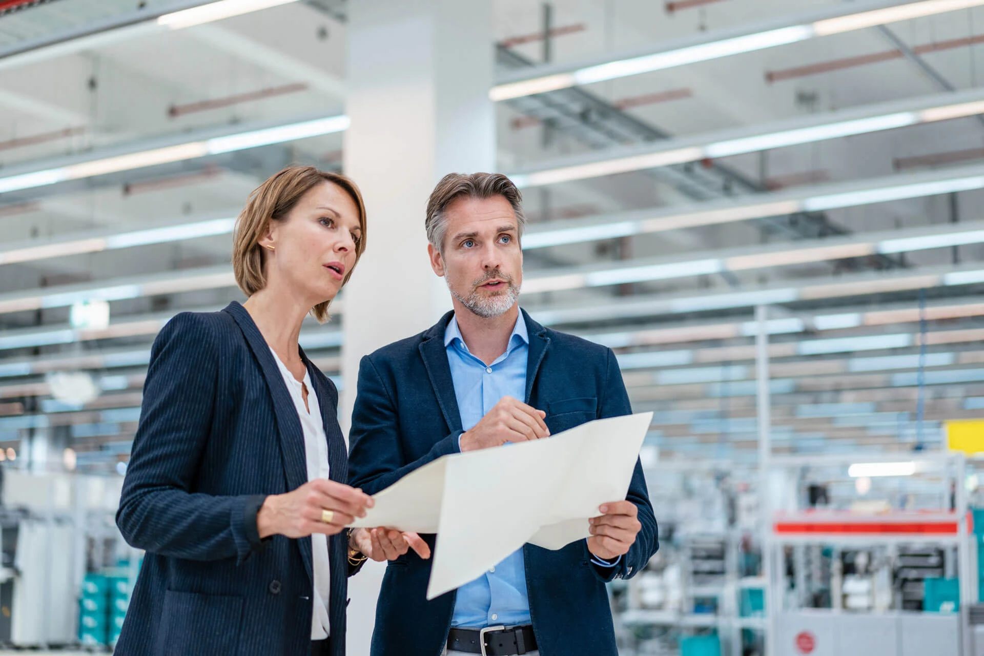 Two professionals in business attire stand in a brightly lit industrial or manufacturing facility, holding and discussing a large blueprint or document, with machinery and equipment visible in the background.