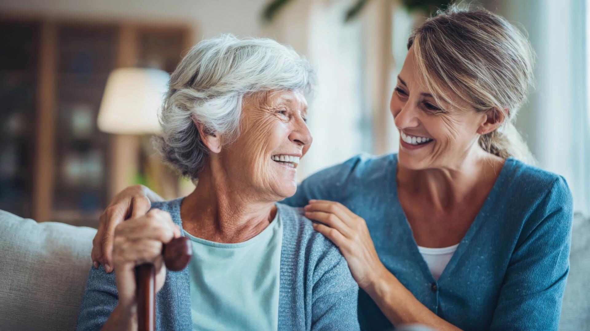 An elderly woman with gray hair and a cane smiles warmly at a younger woman sitting beside her. Both women are laughing and looking at each other, sharing a joyful and affectionate moment indoors.