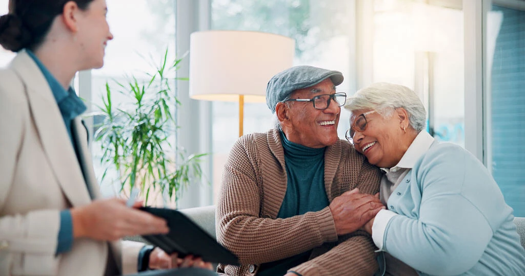 An elderly couple sits close together, smiling and holding hands, while a woman nearby holds a clipboard and smiles at them in a bright, comfortable room.
