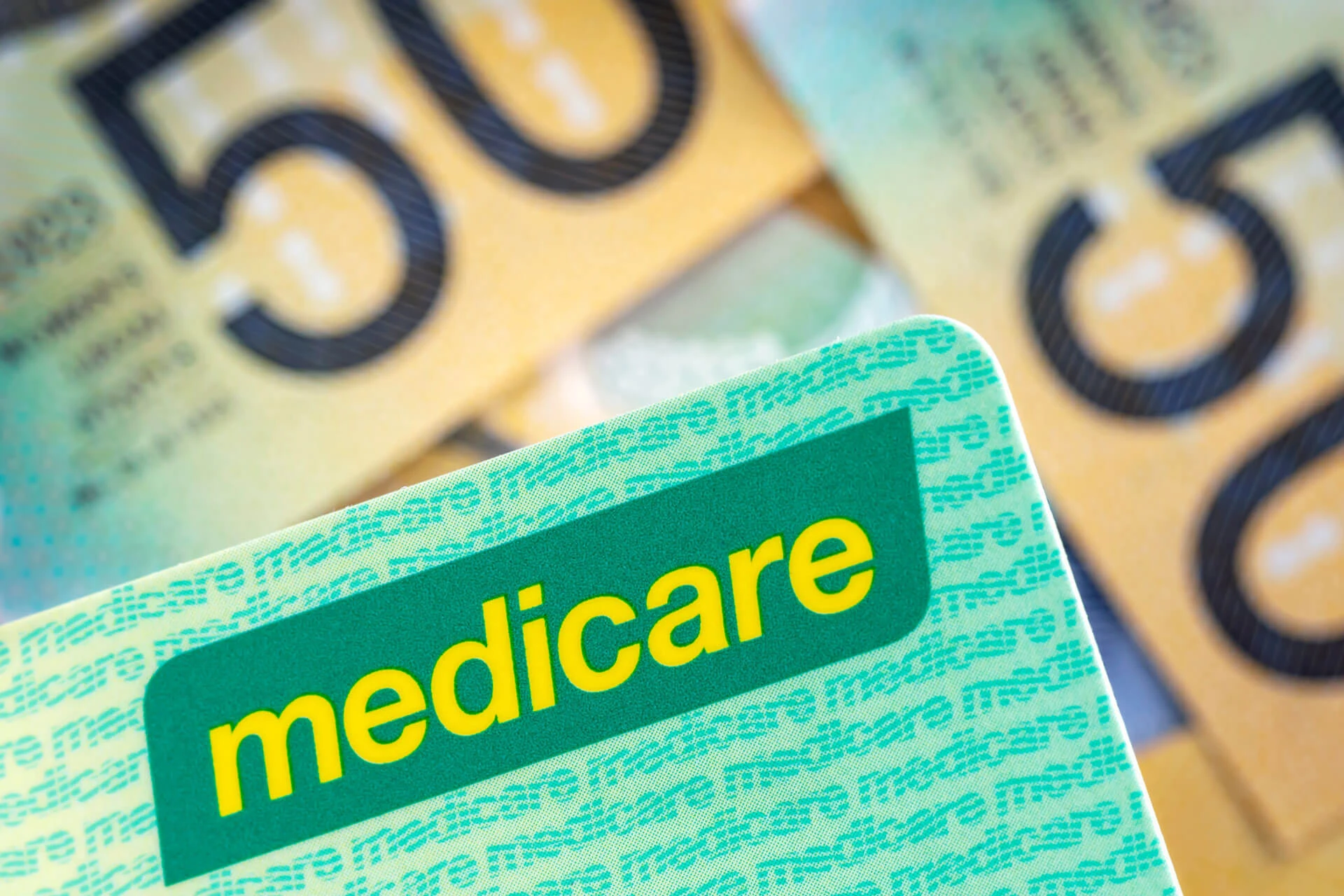 A close-up of an Australian Medicare card partially covering several Australian fifty-dollar banknotes in the background.