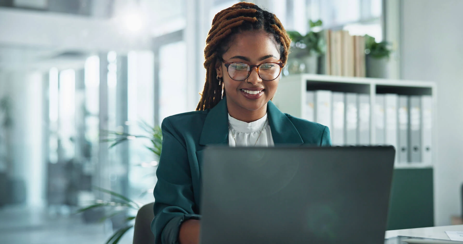 A woman with glasses and long braids is smiling while working on a laptop in a modern office with shelves, folders, and plants in the background.