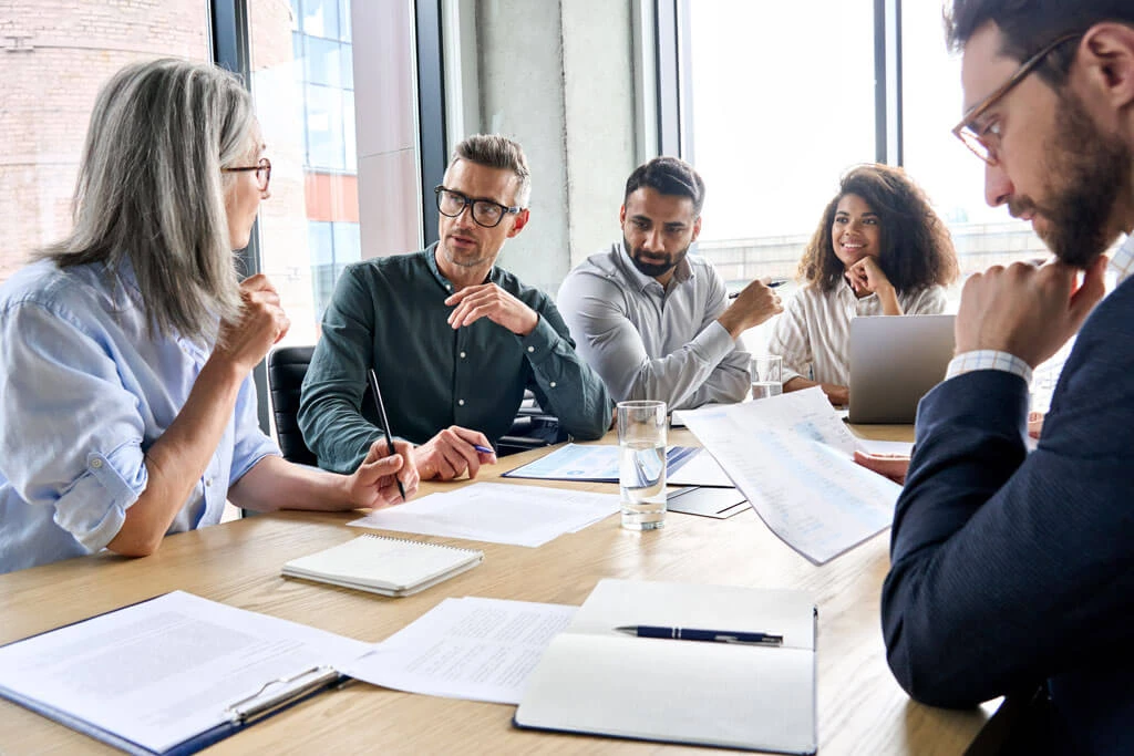 A diverse group of five people sit at a table in a modern office, having a discussion with papers, notebooks, and a laptop in front of them. They appear engaged and attentive.